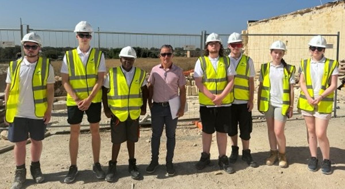 A group of students  in hi-vis vests and hard hats on a sunny construction site, with a group leader